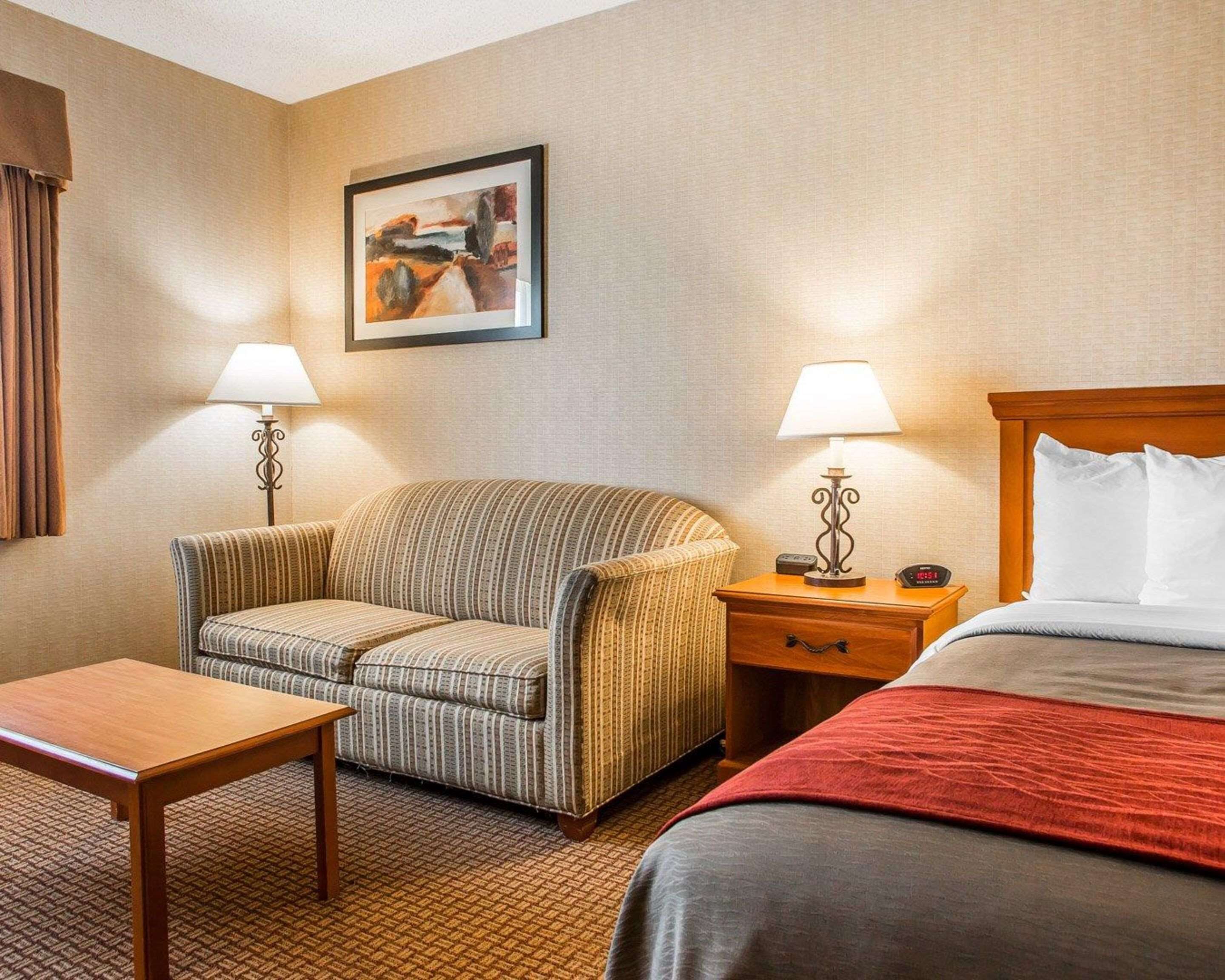 A hotel room with a striped armchair, a wooden nightstand with a lamp, a bed with a red blanket, and a framed painting on the wall.
