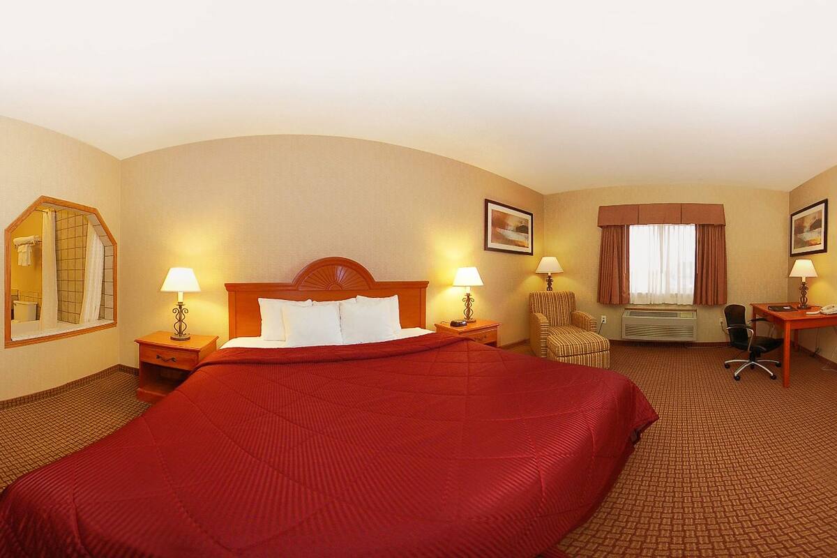 Habitación de hotel con una cama grande, dos mesitas de noche con lámparas, una silla, un escritorio con silla, una ventana con cortinas y un espejo en la pared.