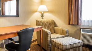 Ett hotellrum med ett skrivbord i trä, en svart stol, en randig fåtölj, en pall, en lampa, en spegel, ett fönster med gardiner och en luftkonditioneringsanläggning.