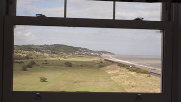 Une vue depuis une fenêtre donnant sur une plage, des champs verdoyants et une zone vallonnée avec des bâtiments.