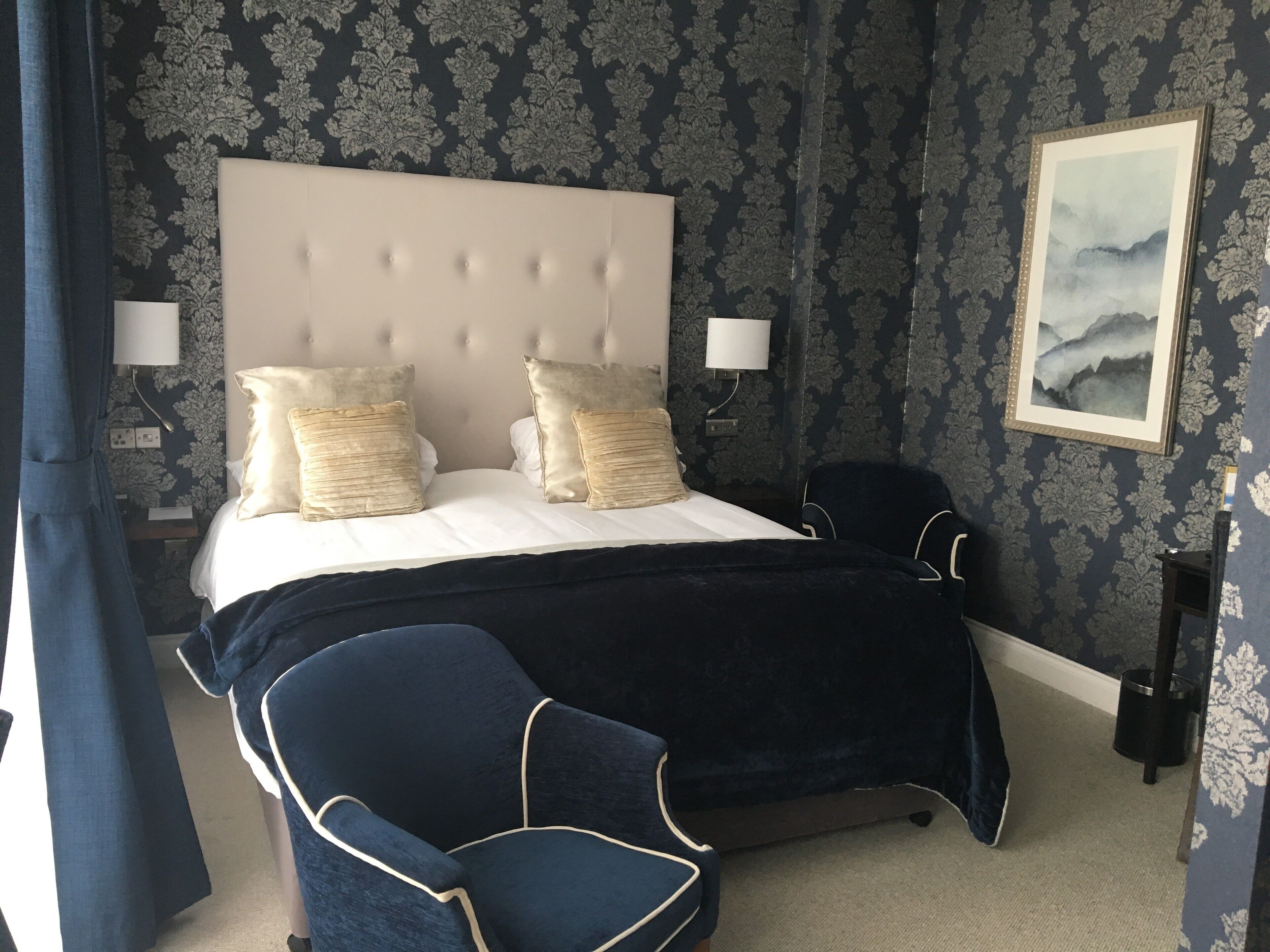 A hotel room with a large bed, two bedside lamps, a chair, and a painting on the wall.