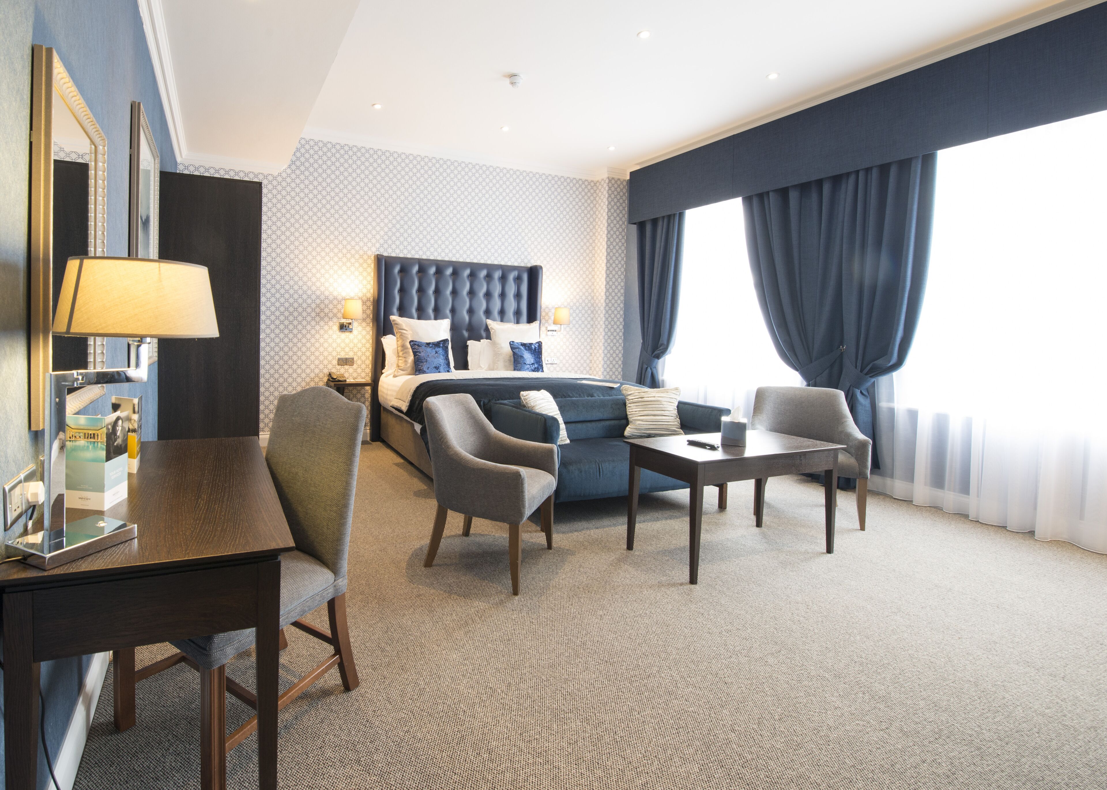 Une chambre d’hôtel comprenant un lit, deux fauteuils, une petite table, un bureau et une lampe.