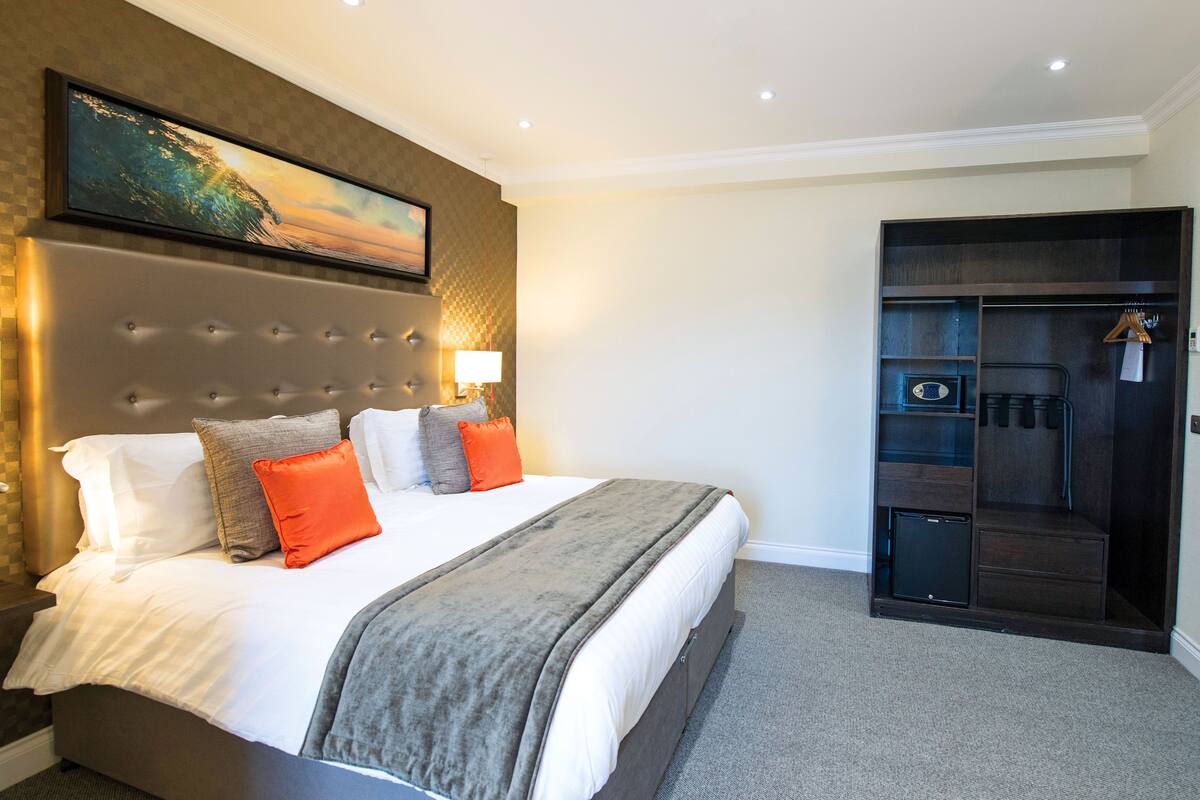 Une chambre d’hôtel avec un grand lit, une tête de lit, un tableau, une armoire et un petit bureau avec une lampe.