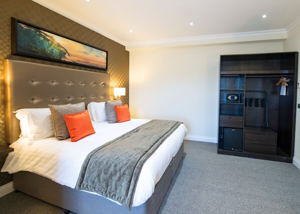 A hotel room with a large bed, a headboard, a painting, a wardrobe, and a small desk with a lamp.