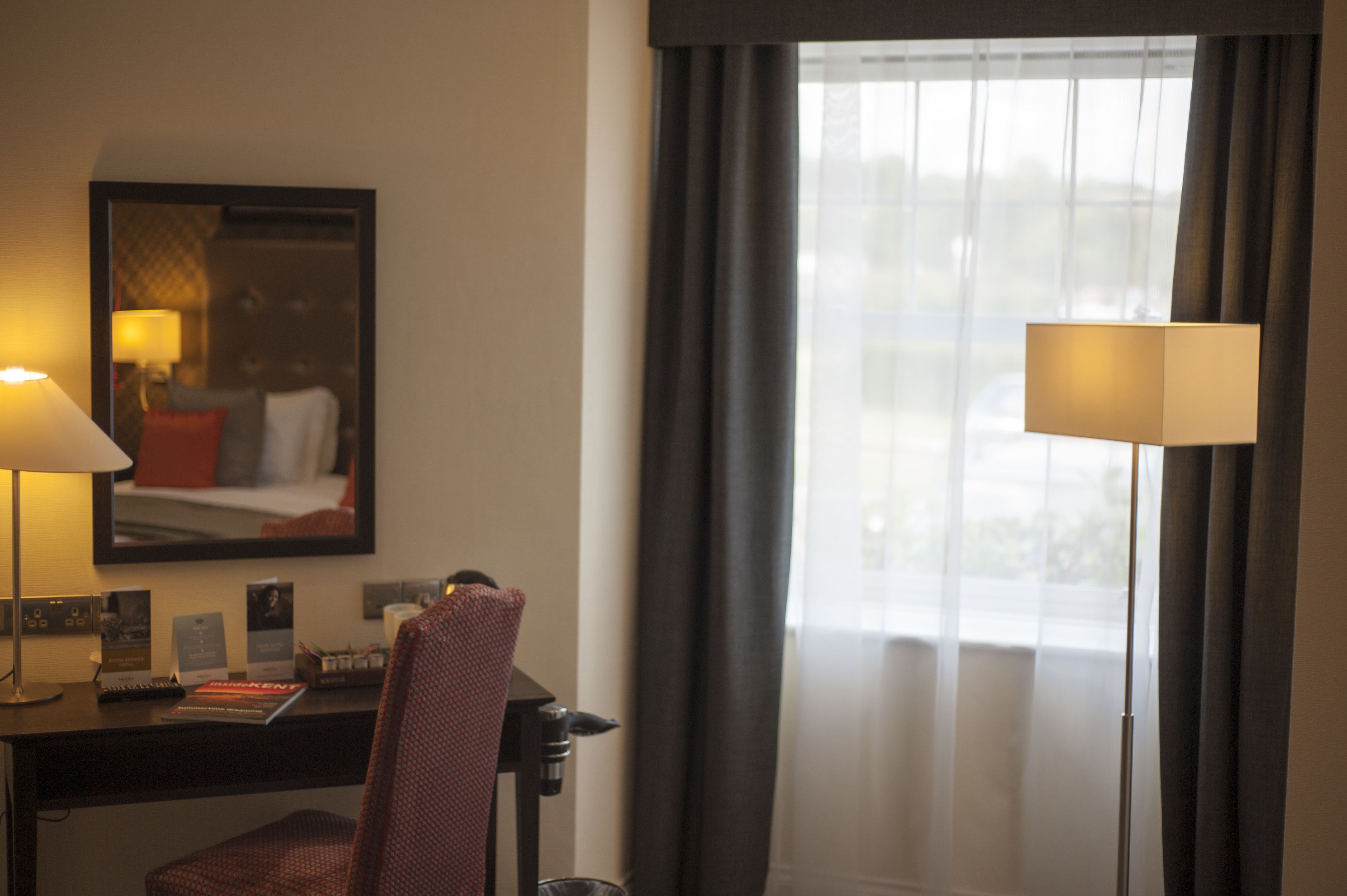 A hotel room with a desk, two lamps, a chair, a bed with a red pillow, a mirror, and a window with curtains.