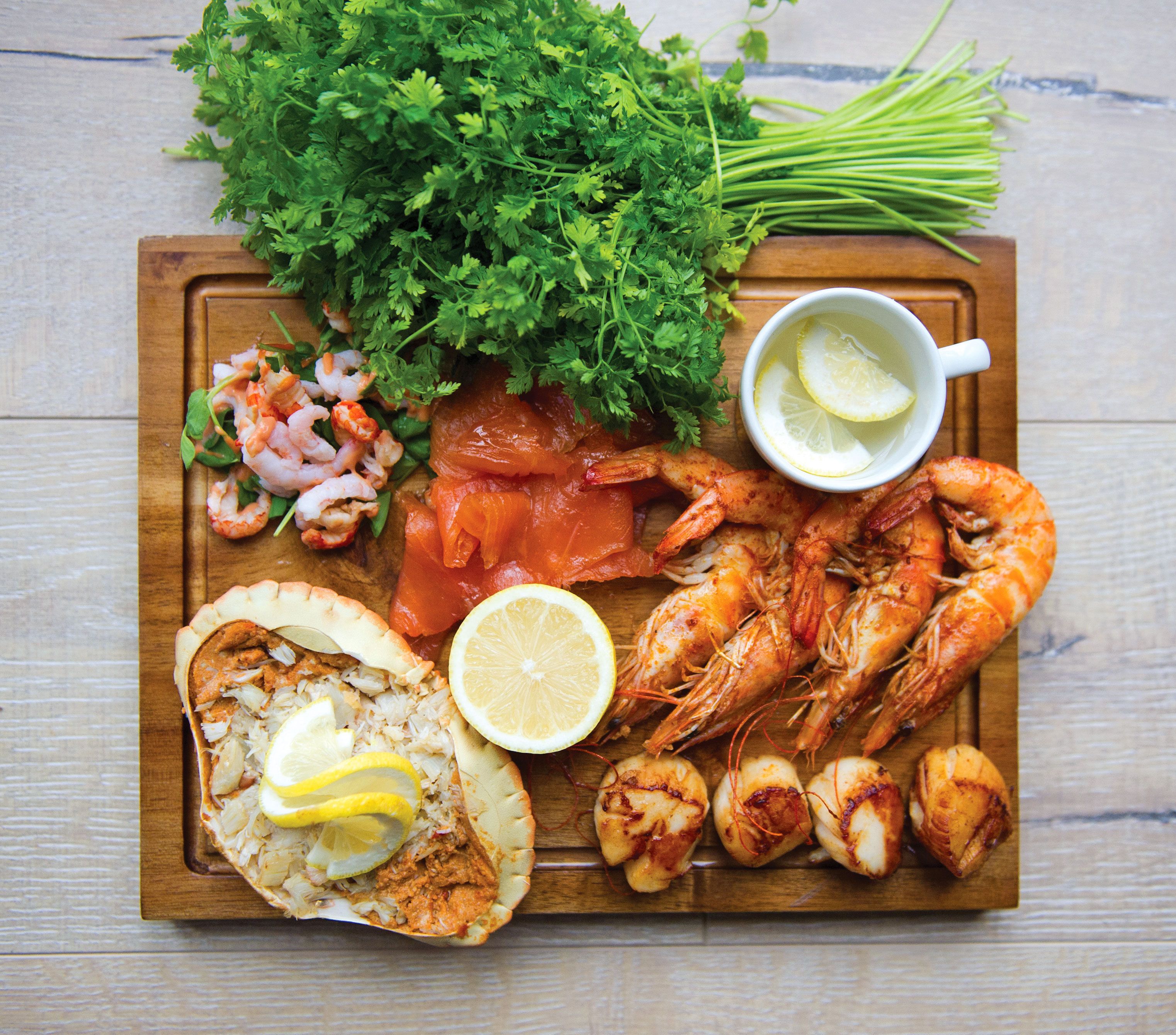 Une planche en bois sur laquelle sont disposés divers fruits de mer, notamment des crevettes, du saumon et des coquilles Saint-Jacques, le tout agrémenté de tranches de citron et de persil.