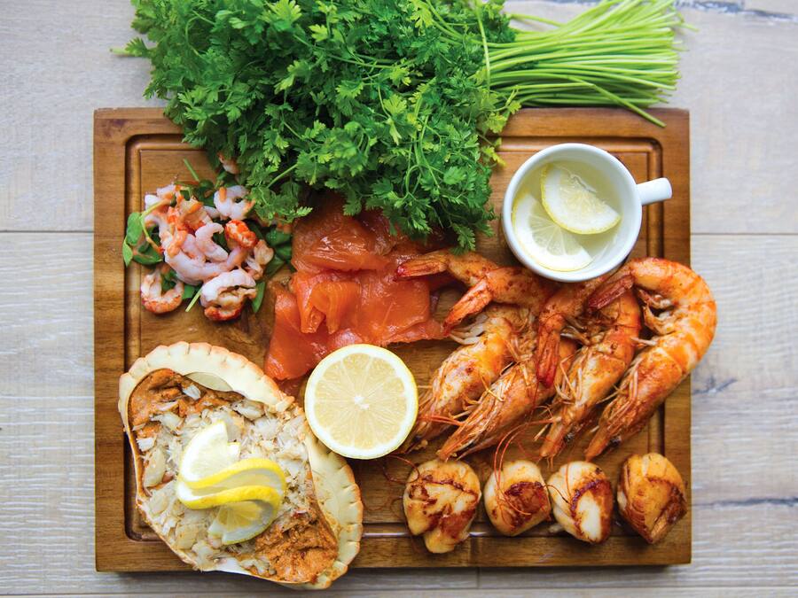 Une planche en bois sur laquelle sont disposés divers fruits de mer, notamment des crevettes, du saumon et des coquilles Saint-Jacques, le tout agrémenté de tranches de citron et de persil.