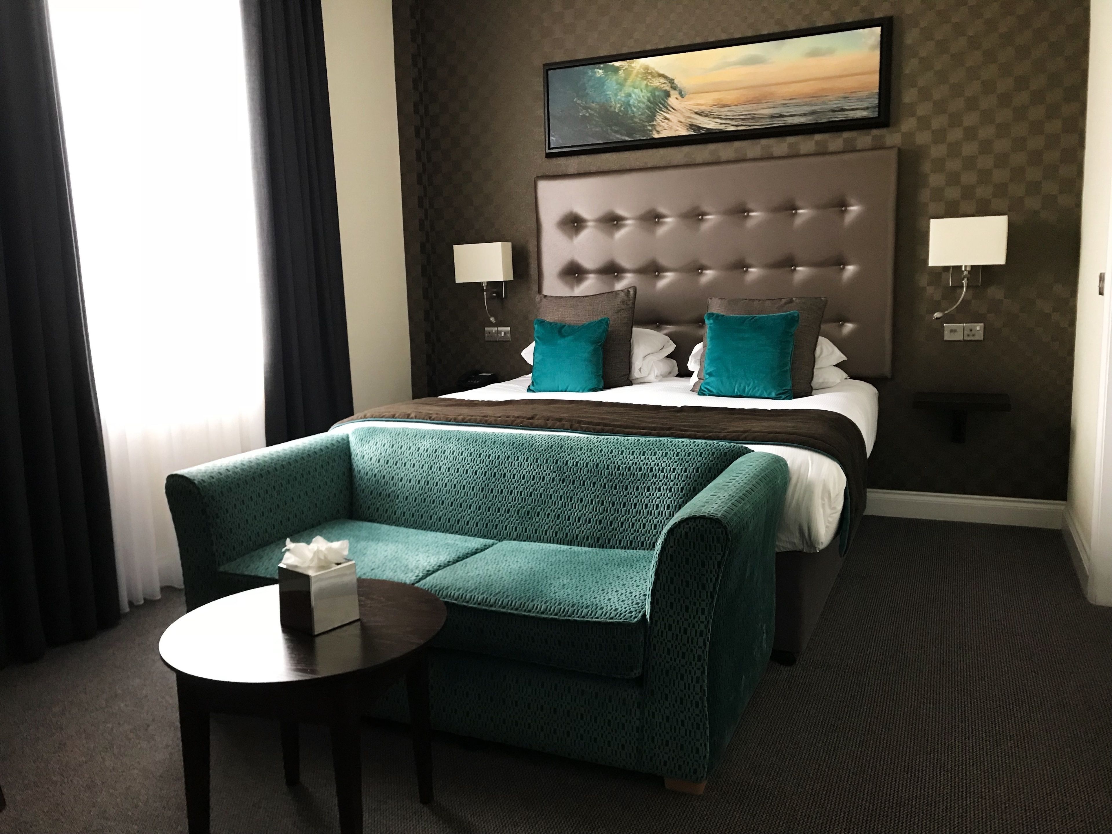 A hotel room with a bed, a green sofa, a small table with a tissue box, a wall-mounted TV, and a painting of a beach scene.