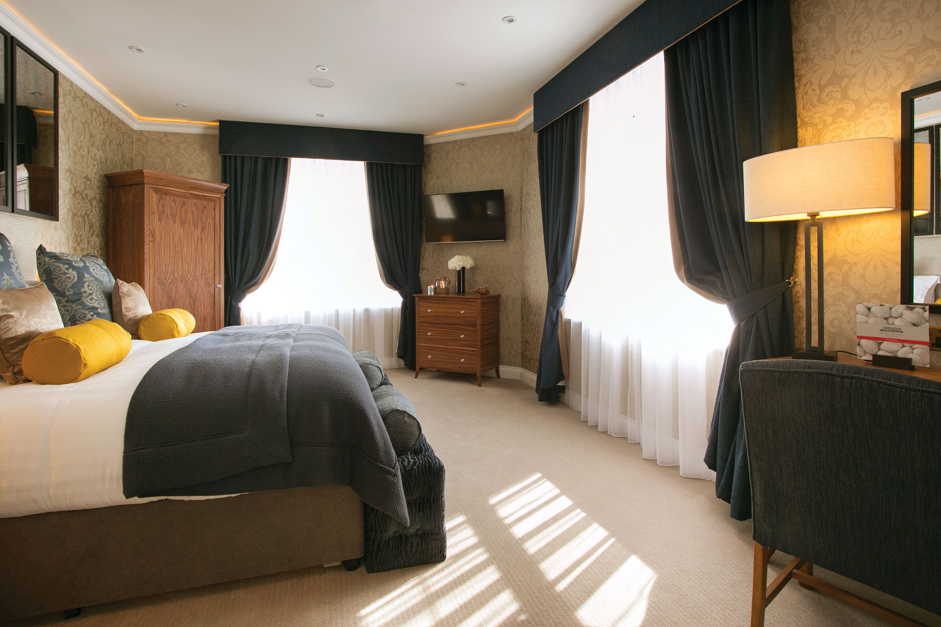 A hotel room with a large bed, a wooden dresser, a desk with a lamp, and a flat-screen TV mounted on the wall.