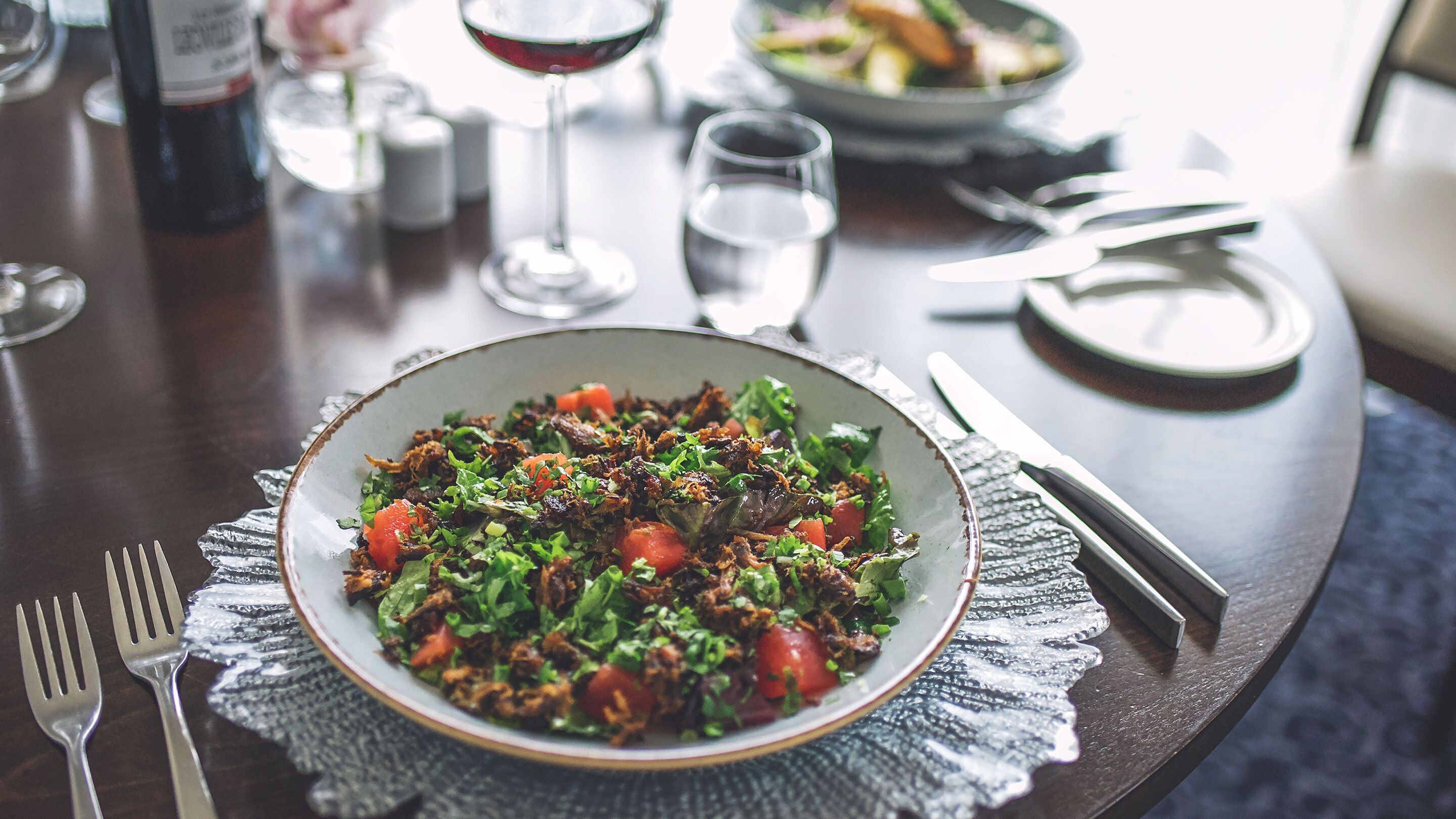 A dining table with a bowl of salad, cutlery, and a wine bottle.
