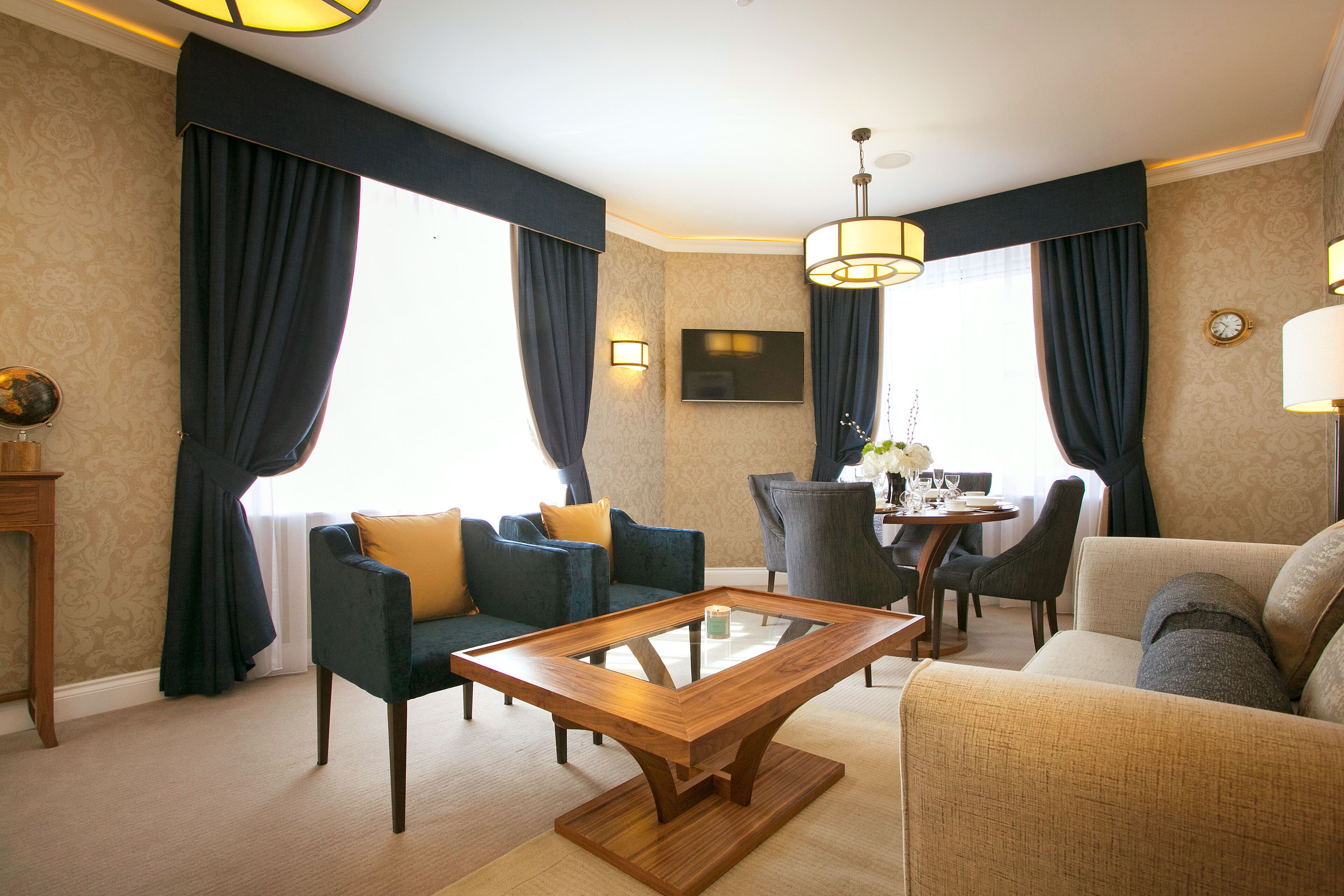 Une chambre d’hôtel moderne avec une table basse en verre, des rideaux bleu foncé, un canapé, un fauteuil et un coin repas comprenant une table et des chaises.