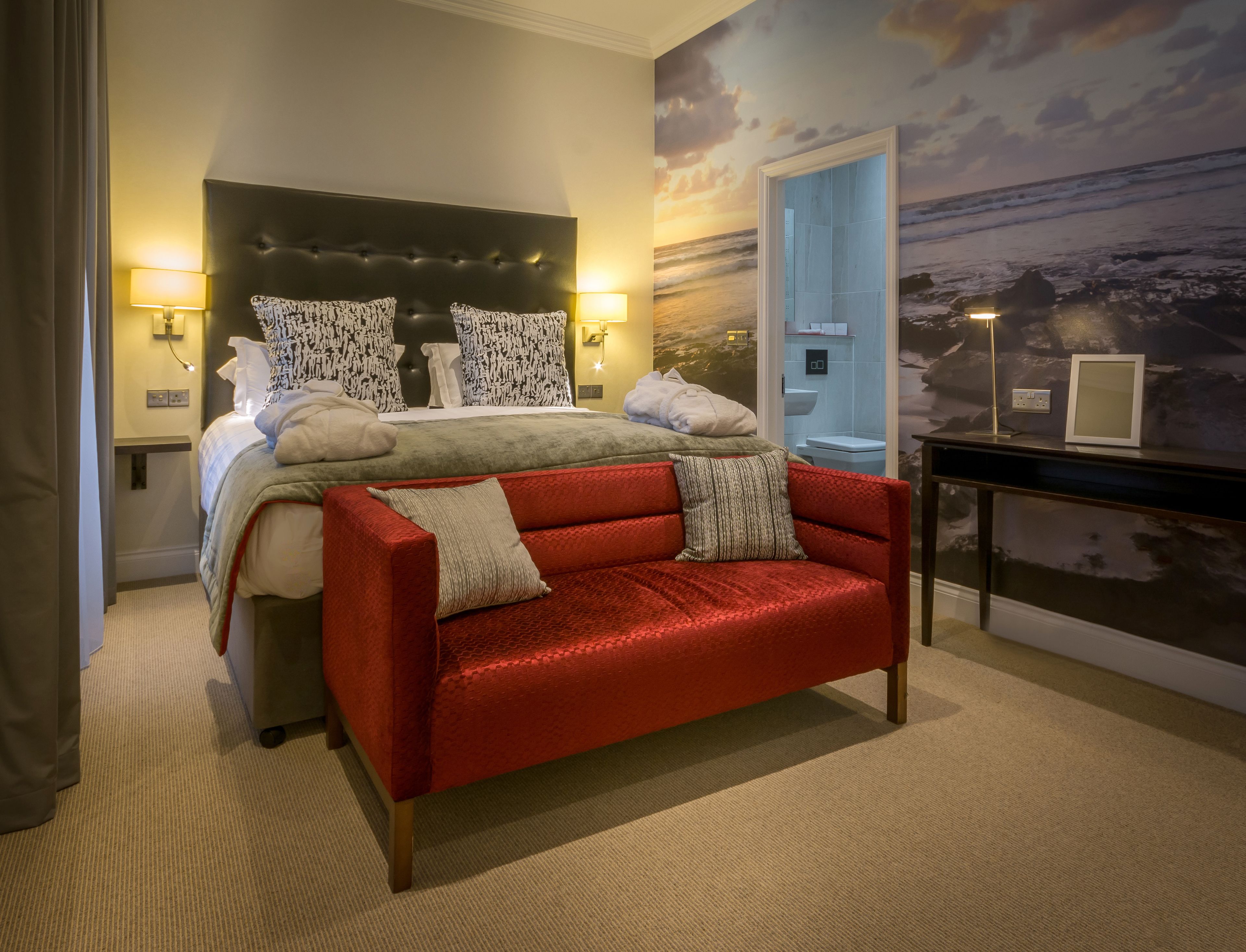 A hotel room with a large bed, a red sofa, a nightstand, and a wall mural of a beach scene.
