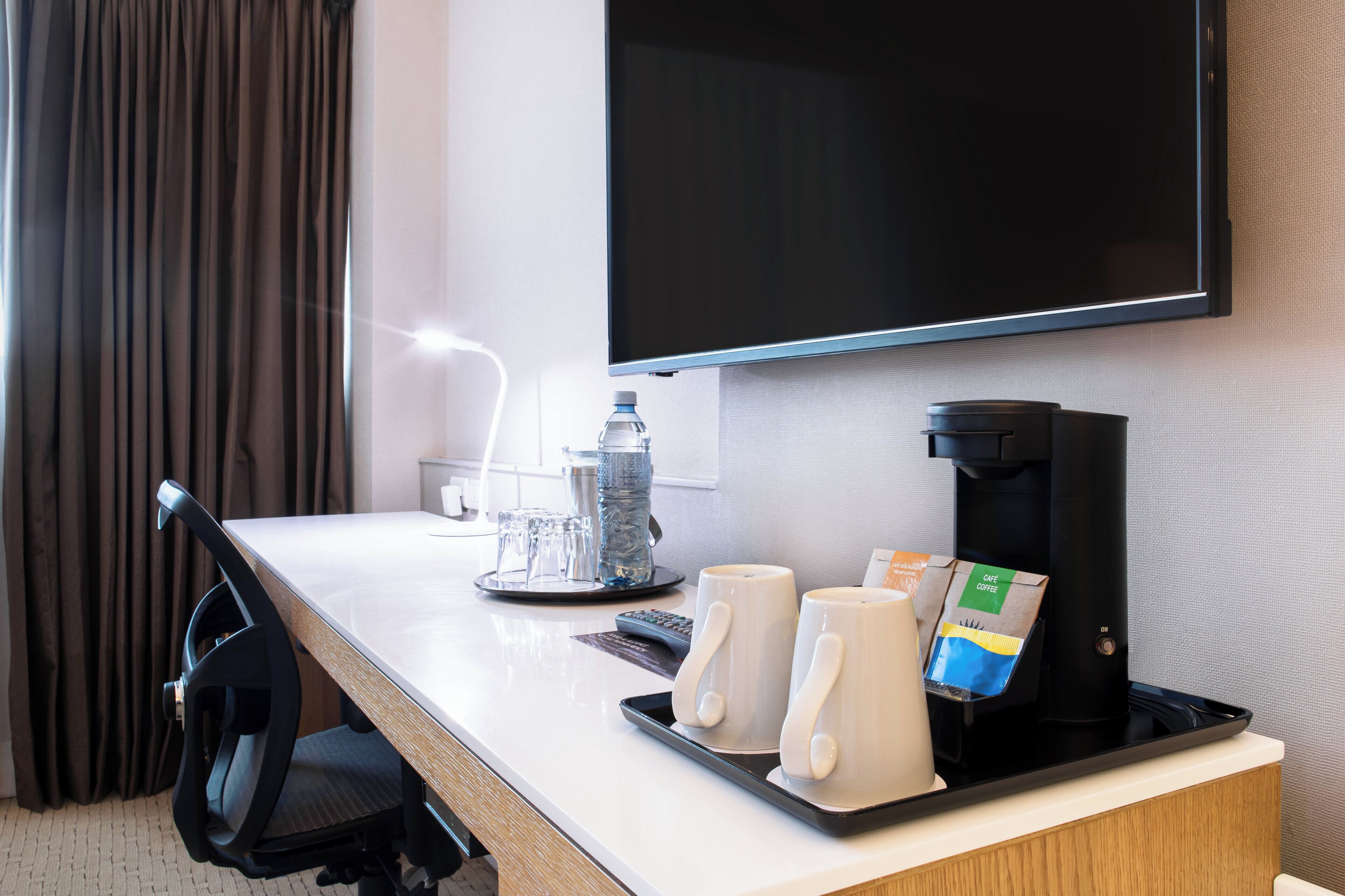 A hotel room with a desk, a coffee maker, two mugs, a bottle of water, and a television.