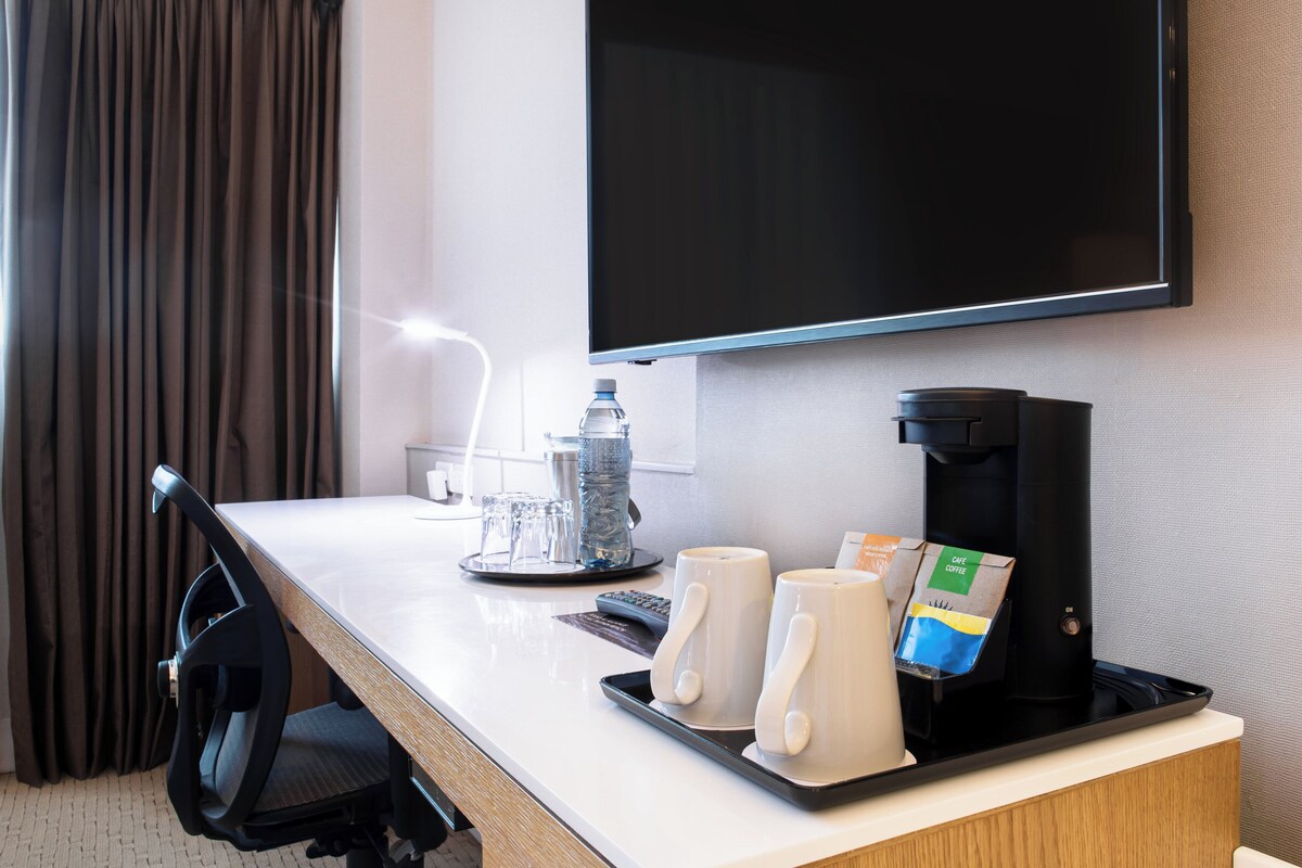 A hotel room with a desk, a coffee maker, two mugs, a bottle of water, and a television.
