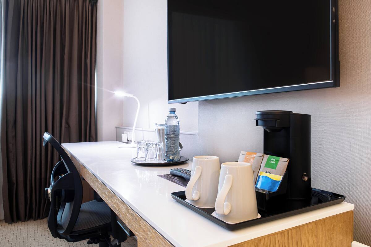 A hotel room with a desk, a coffee maker, two mugs, a bottle of water, and a television.
