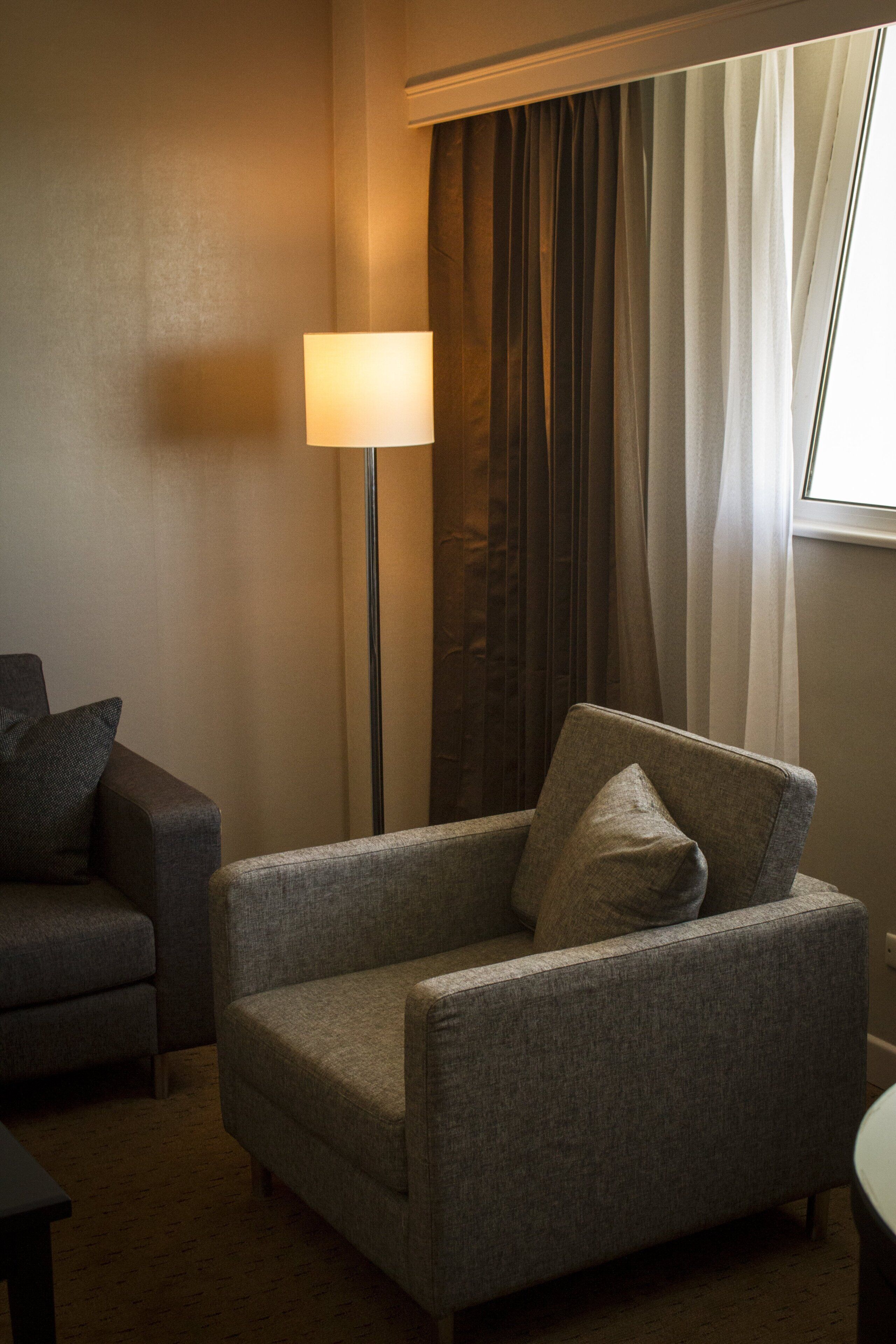 A hotel room with a grey armchair, a floor lamp, and a window with curtains.