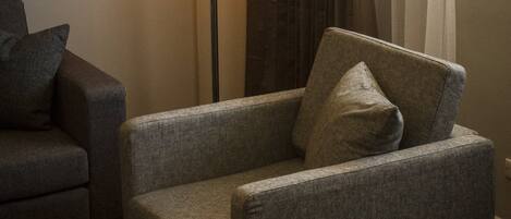 A hotel room with a grey armchair, a floor lamp, and a window with curtains.