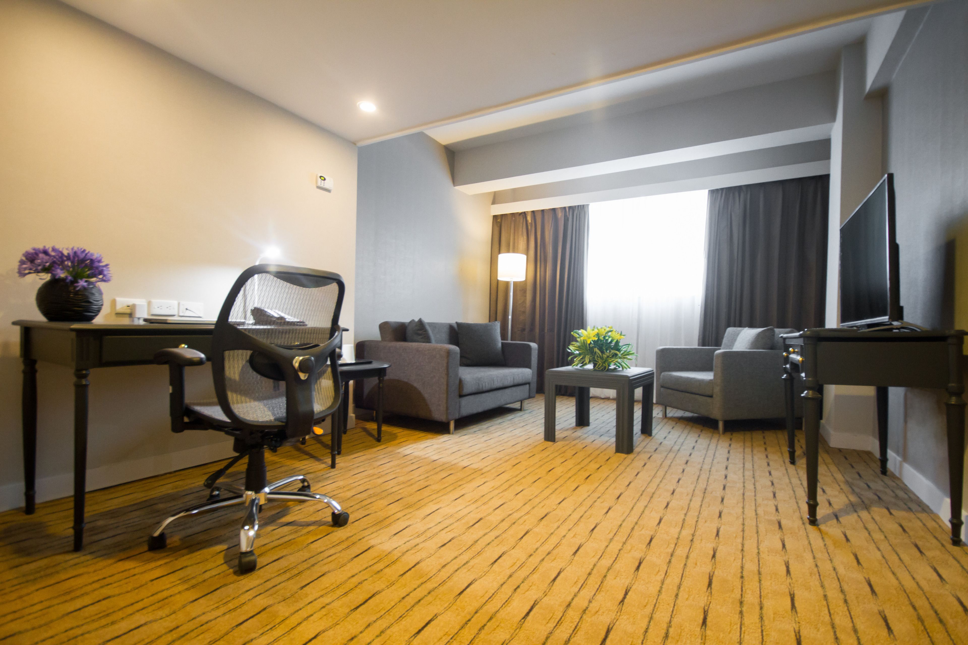 Una camera d'albergo con pavimento in legno, una scrivania con un computer, due divani grigi, un tavolino con una pianta e una TV su un mobile.