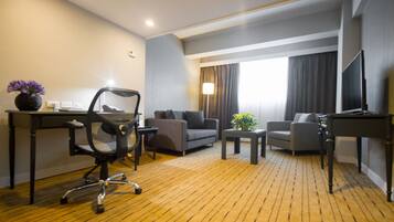 A hotel room with a wooden floor, a desk with a computer, two grey sofas, a small table with a plant, and a TV on a stand.