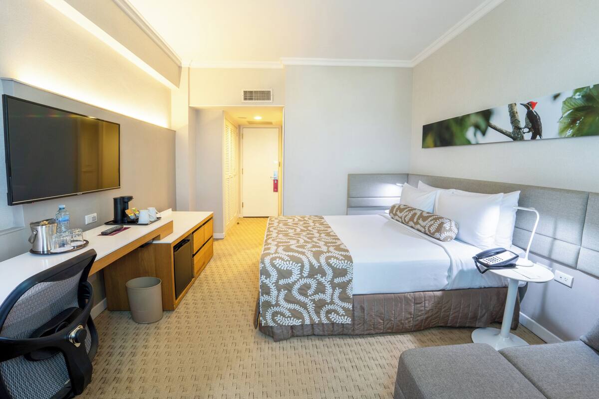 A hotel room with a bed, a desk with a chair, a TV, a painting on the wall, and a small table with a lamp.