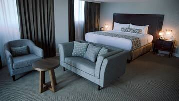 A hotel room with a bed, a sofa, a chair, a small table, and a nightstand with a lamp.