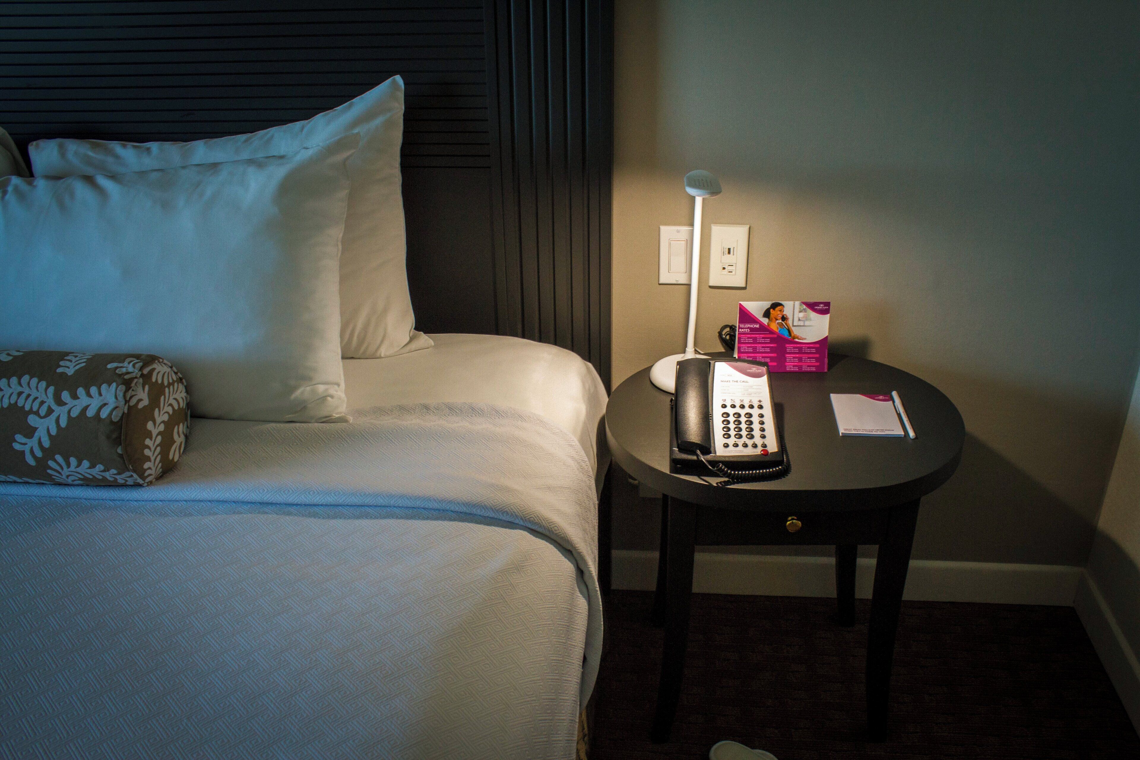 Habitación de hotel con una cama, una mesita de noche con un teléfono y un folleto, y un enchufe en la pared.