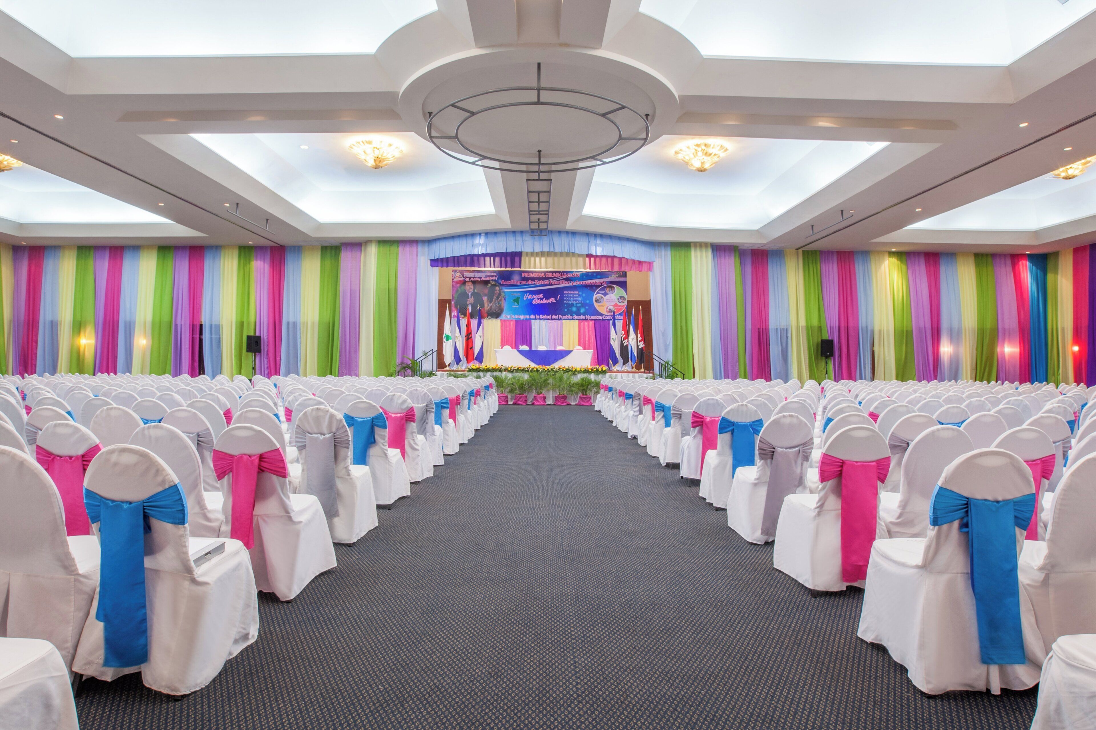 A conference room with rows of chairs, a stage with a backdrop, and a high ceiling with skylights.