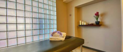 A room with a bench, a window with a grid pattern, and a shelf with candles and flowers.