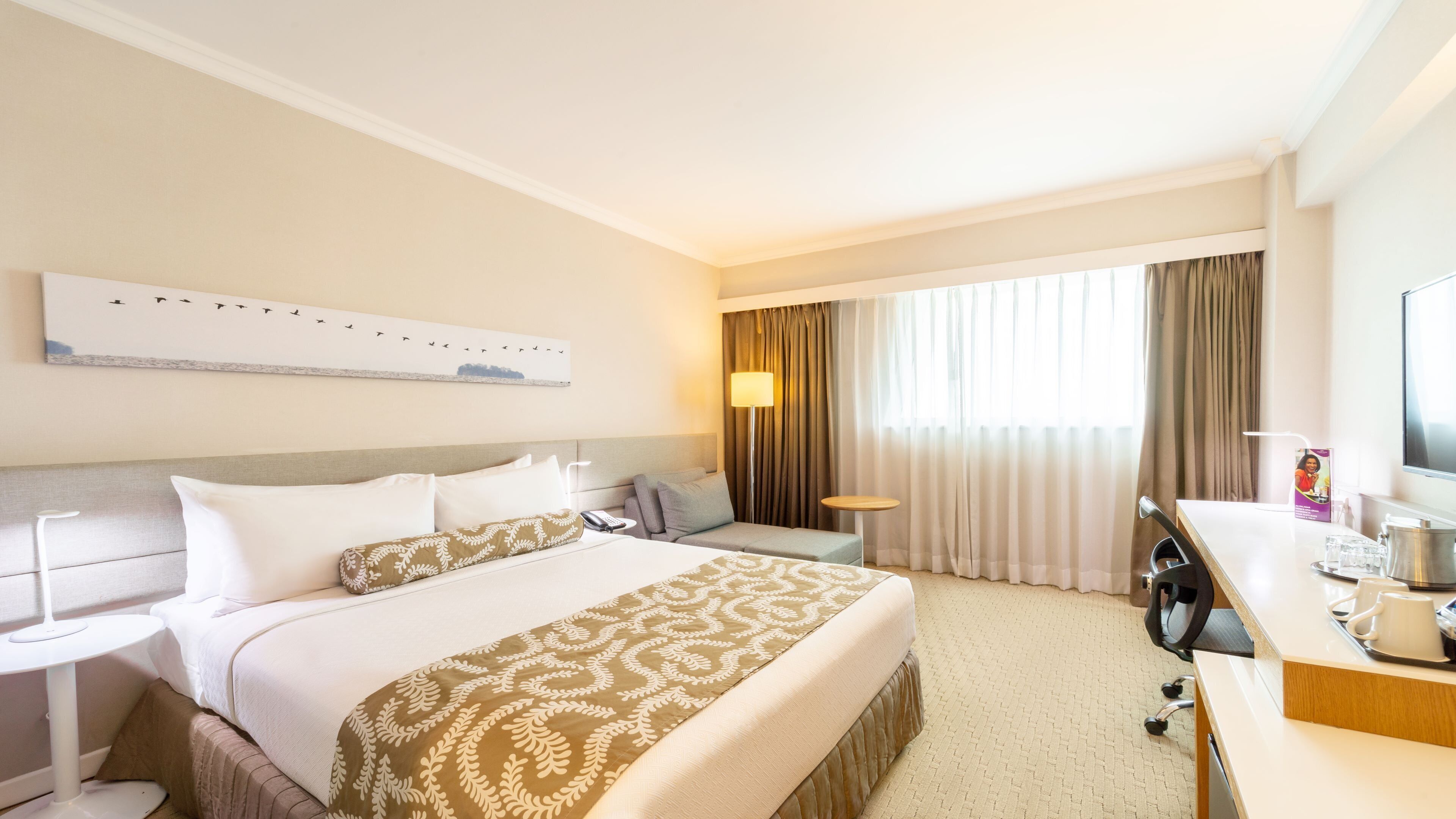A hotel room with a bed, bedside tables, a desk, a chair, a lamp, a television, and curtains.
