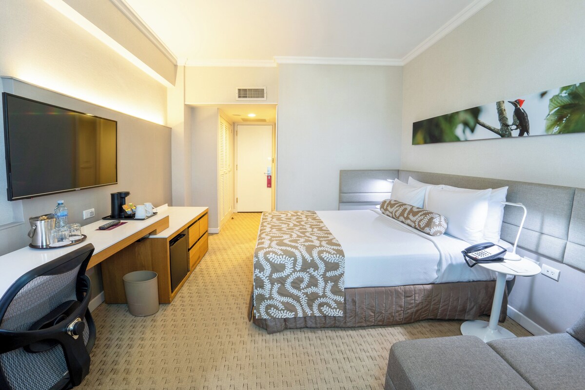 A hotel room with a bed, a desk with a chair, a TV, a painting on the wall, and a small table with a lamp.