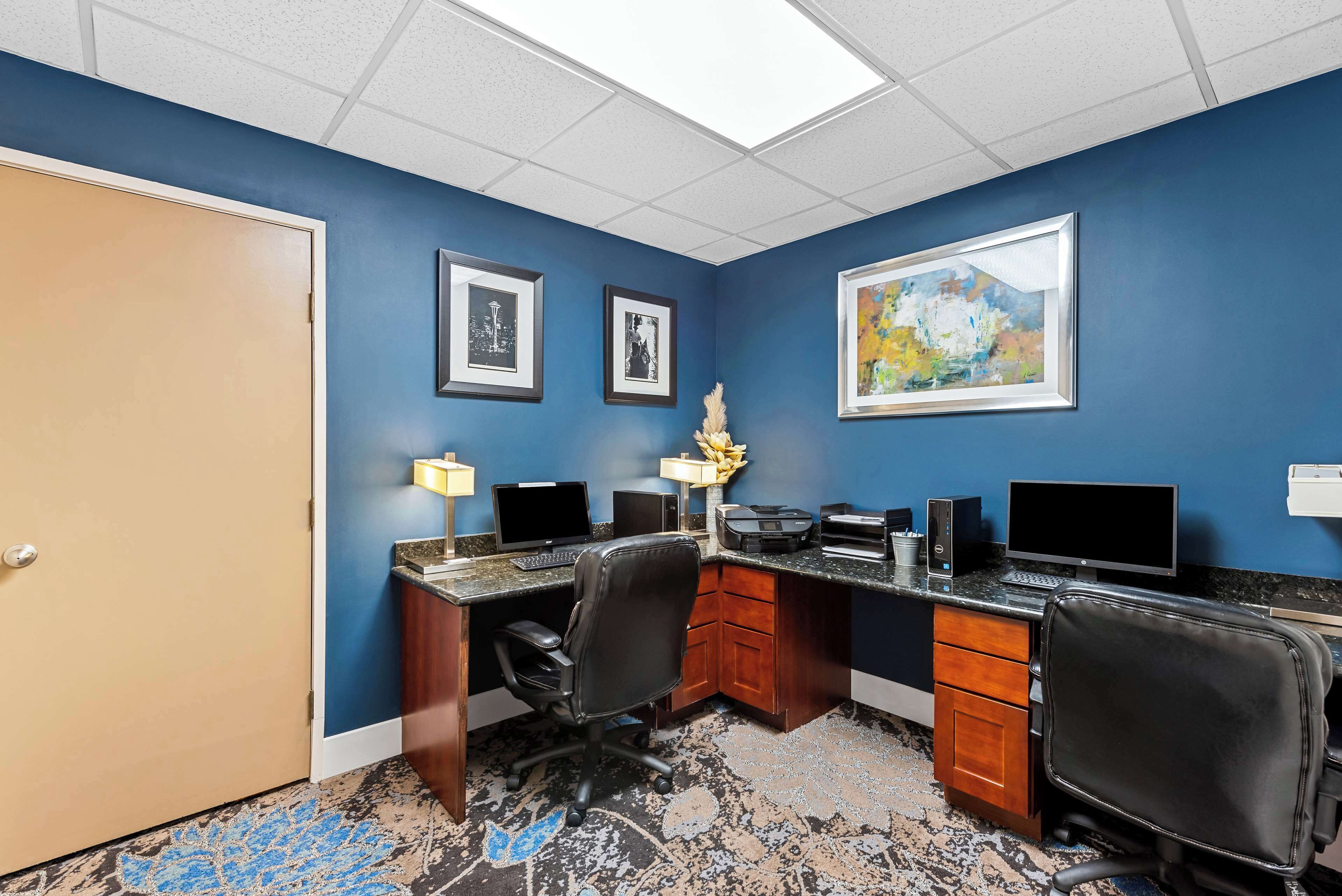 A conference room with a blue wall, a wooden desk, two chairs, a computer, and framed pictures.