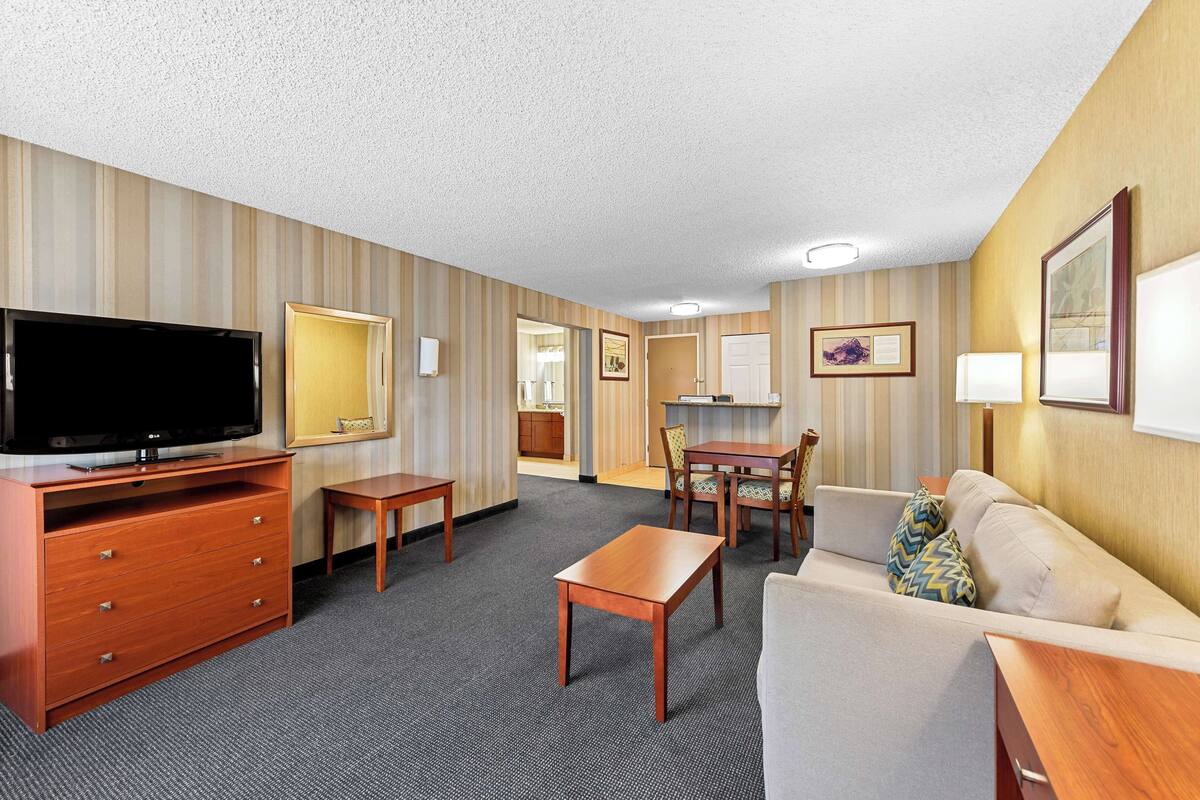A hotel room with a flat-screen TV, wooden furniture, a sofa, a dining table, and a kitchen area.