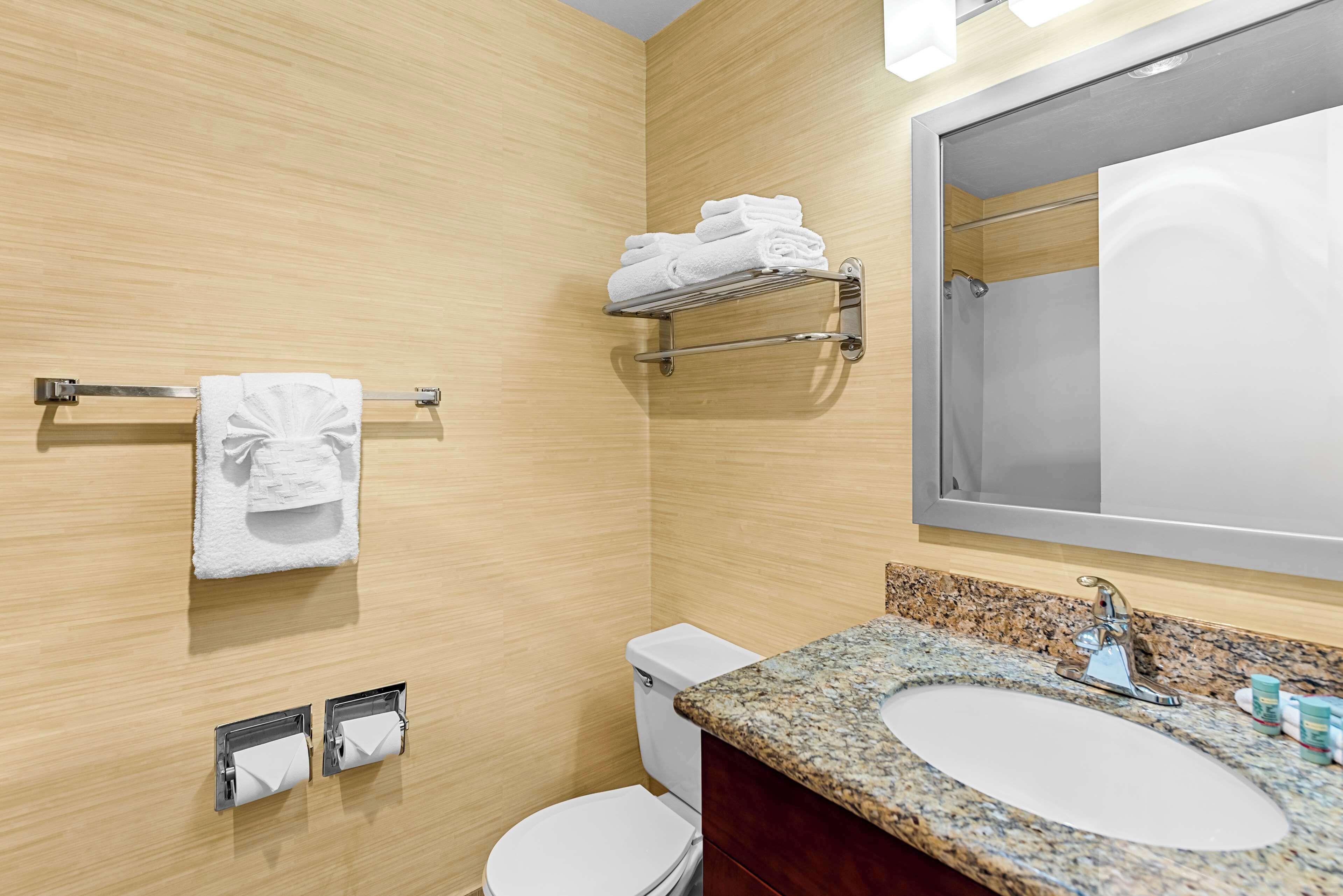 A modern bathroom with a wooden wall, a sink with a granite countertop, a toilet, a towel rack with folded towels, and a mirror.