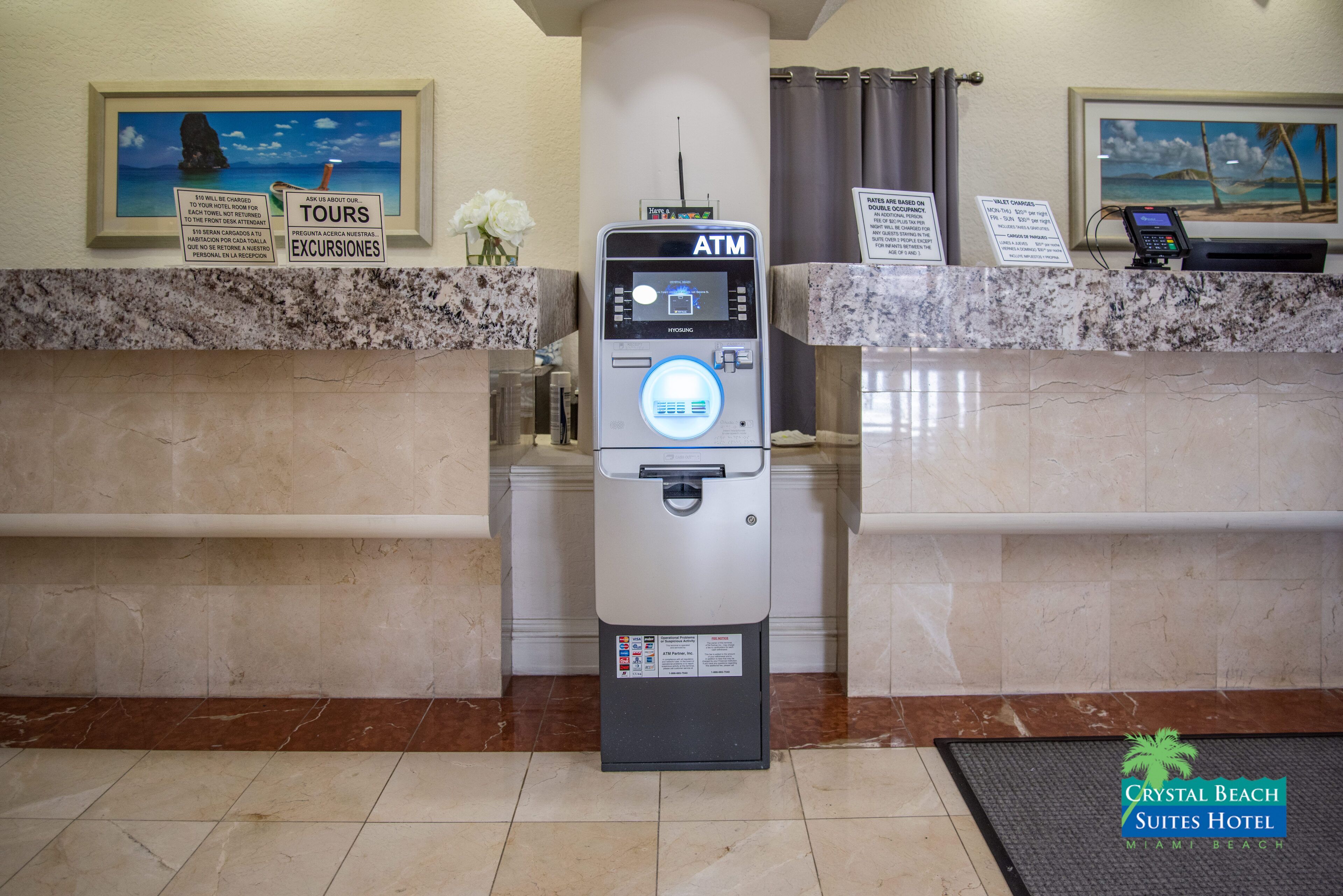An ATM machine at a hotel reception with a marble counter, a framed picture of a beach, and a sign for tours and excursions.