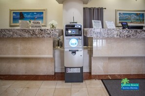 An ATM machine at a hotel reception with a marble counter, a framed picture of a beach, and a sign for tours and excursions.