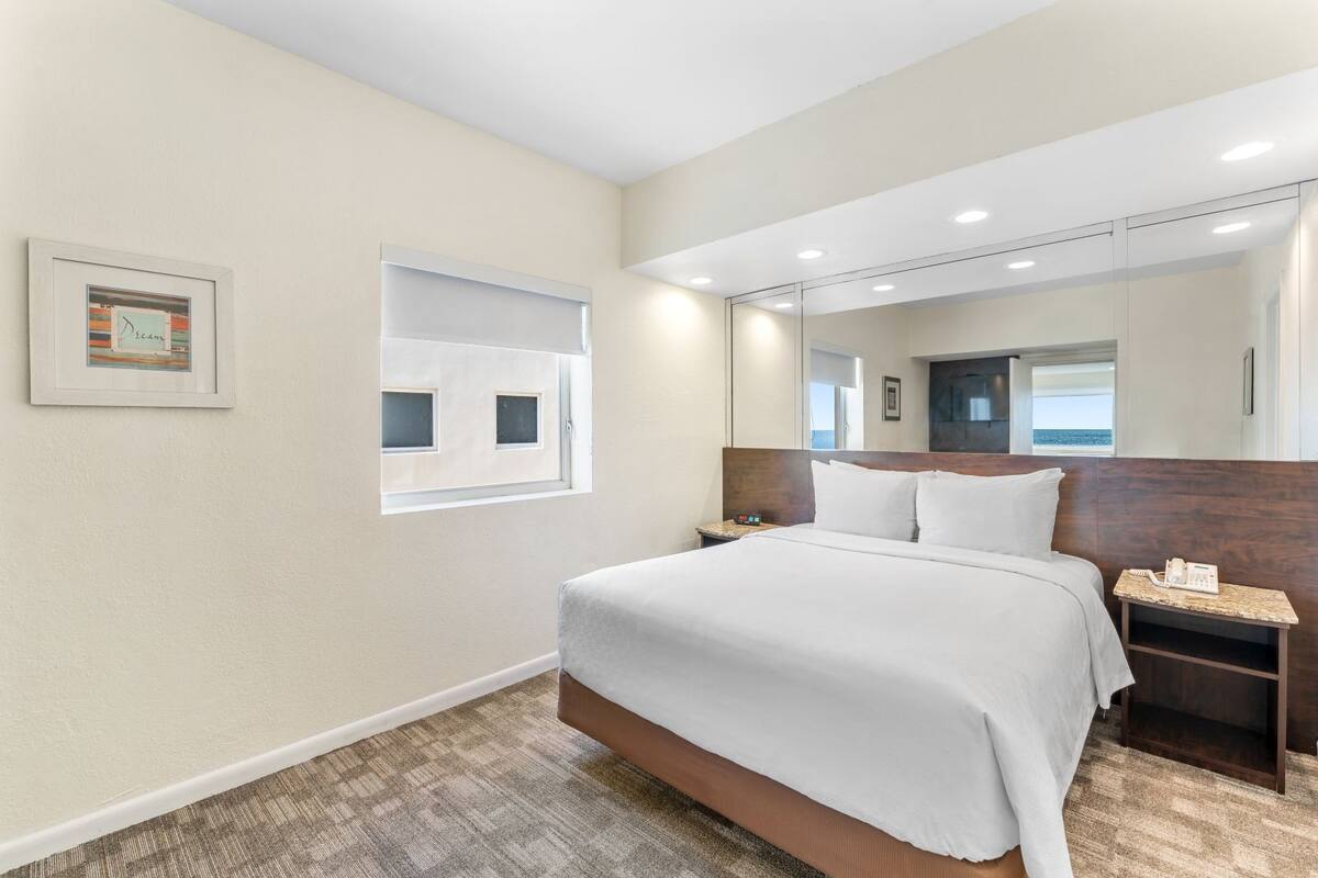 A hotel room with a large bed, a bedside table with a phone, a window, and a framed picture on the wall.