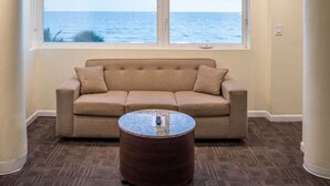 A room with a beige sofa, a round coffee table, and a large window overlooking the ocean.