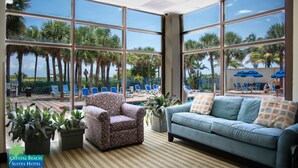 A hotel room with large windows, a patterned armchair, a blue sofa, and a view of a pool area with palm trees.