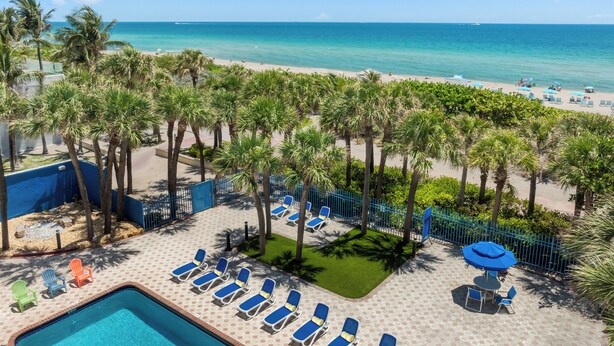 A pool area with lounge chairs and a pool, surrounded by palm trees and a beach view.