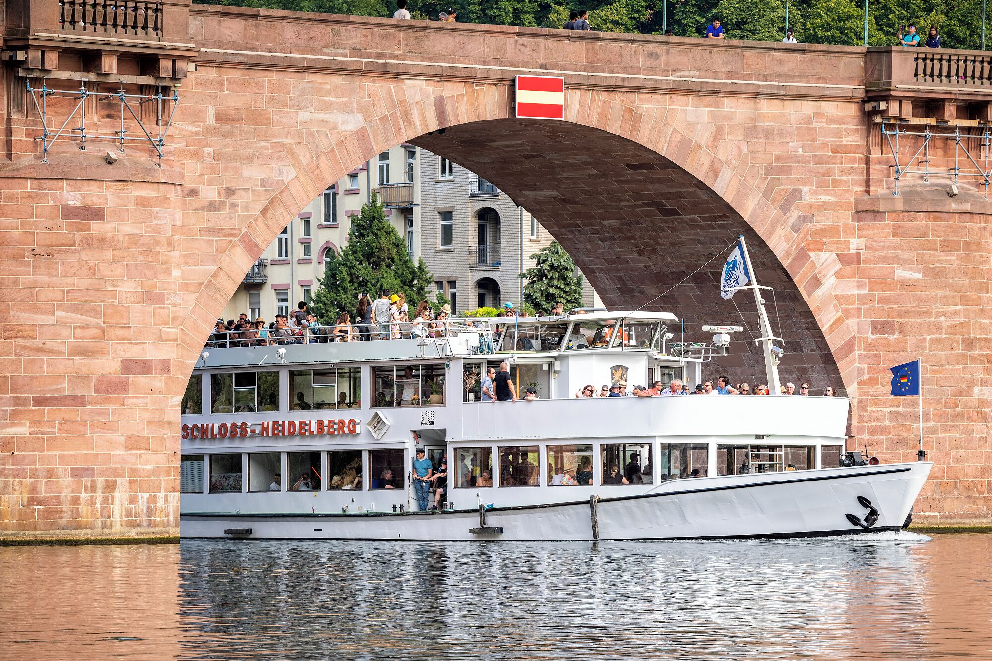 A white boat with a restaurant deck named "SCHLOSS-HEIDELBERG" docked under a stone bridge.