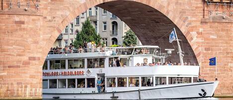 A white boat with a restaurant deck named "SCHLOSS-HEIDELBERG" docked under a stone bridge.