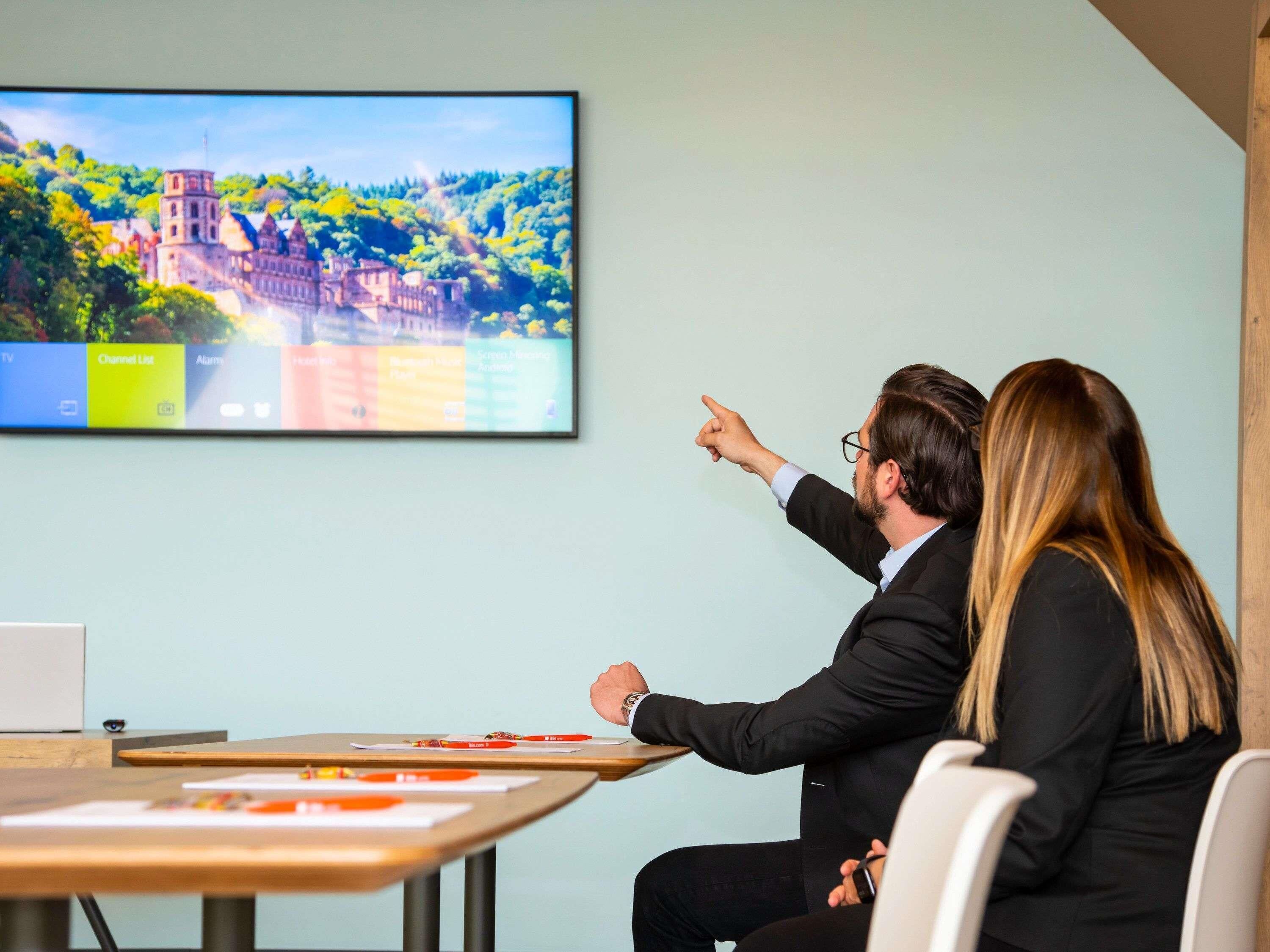 Une salle de conférence dotée d’un grand écran diffusant un paysage pittoresque, d’une table en bois sur laquelle sont disposés des documents, et où deux personnes sont engagées dans une discussion.