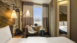 A hotel room with a large bed, a desk with a chair, a telephone, a hat, and a suitcase. - ibis Heidelberg Hauptbahnhof (Heidelberg)