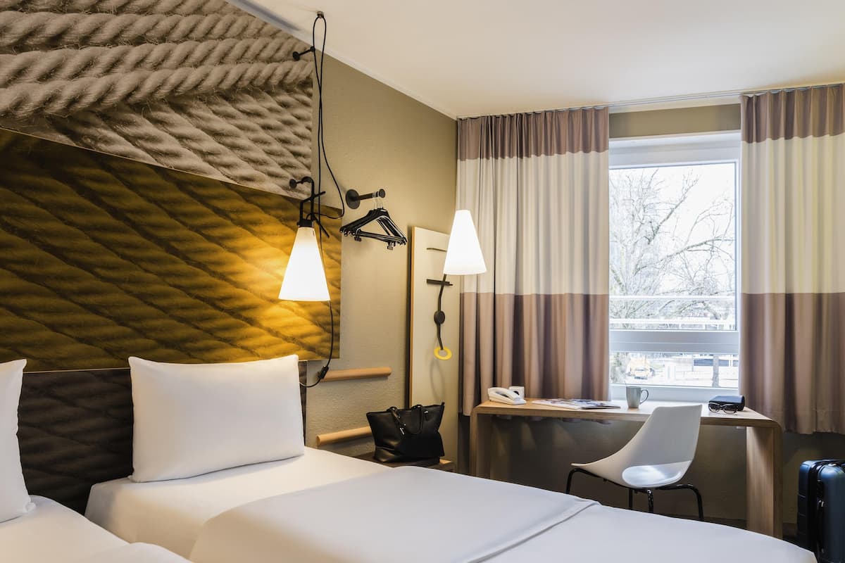 Une chambre d’hôtel avec deux lits, un bureau, une chaise, une lampe, un téléphone et une fenêtre avec des rideaux.