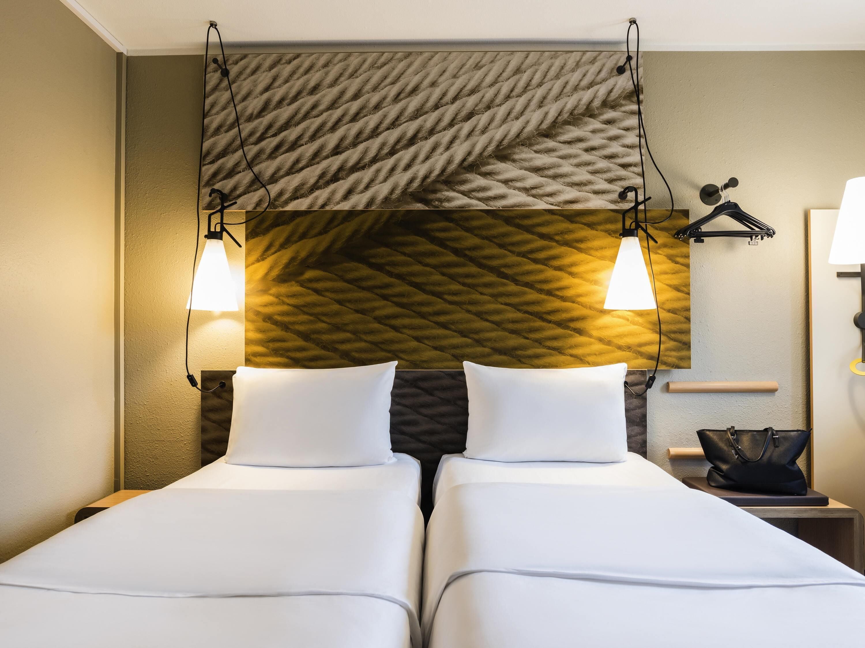 Ein Hotelzimmer mit zwei Betten, einer strukturierten Wand, Wandleuchten, einem kleinen Tisch mit Lampe und einem hängenden Kleiderständer.