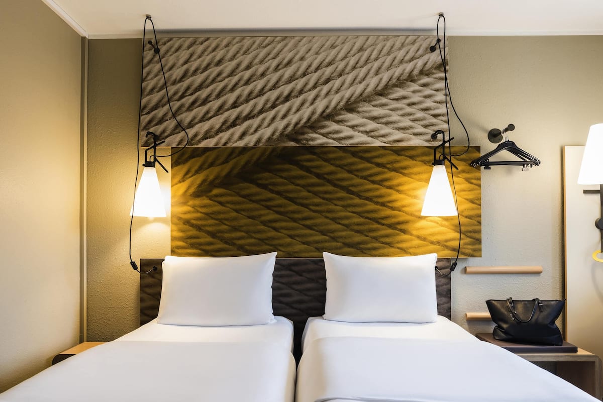 Ein Hotelzimmer mit zwei Betten, einer strukturierten Wand, Wandleuchten, einem kleinen Tisch mit Lampe und einem hängenden Kleiderständer.