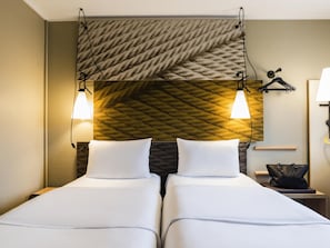 Une chambre dâhĂŽtel avec deux lits, un mur Ă texture, des lampes fixĂ©es au mur, une petite table avec une lampe et un porte-manteau suspendu.