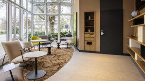 A modern interior with large windows, a wooden bookshelf, and a seating area with tables and chairs. - ibis Heidelberg Hauptbahnhof (Heidelberg)