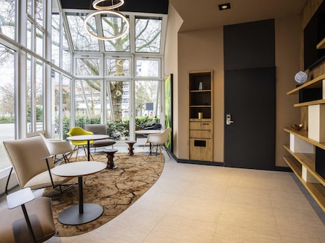 A modern interior with large windows, a wooden bookshelf, and a seating area with tables and chairs.. ibis Heidelberg Hauptbahnhof