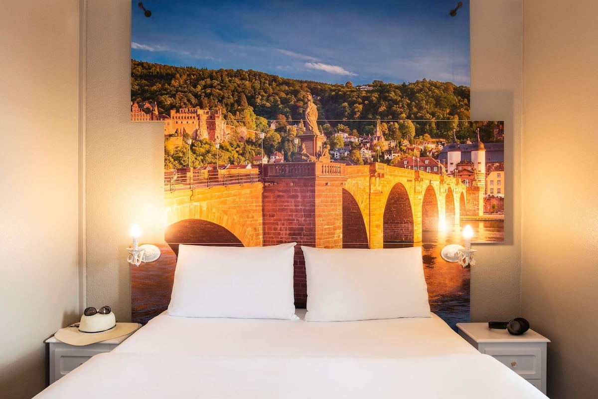 Ein Hotelzimmer mit einem Bett, zwei Nachttischen, einer Lampe und einem großen Wandbild einer Brücke.